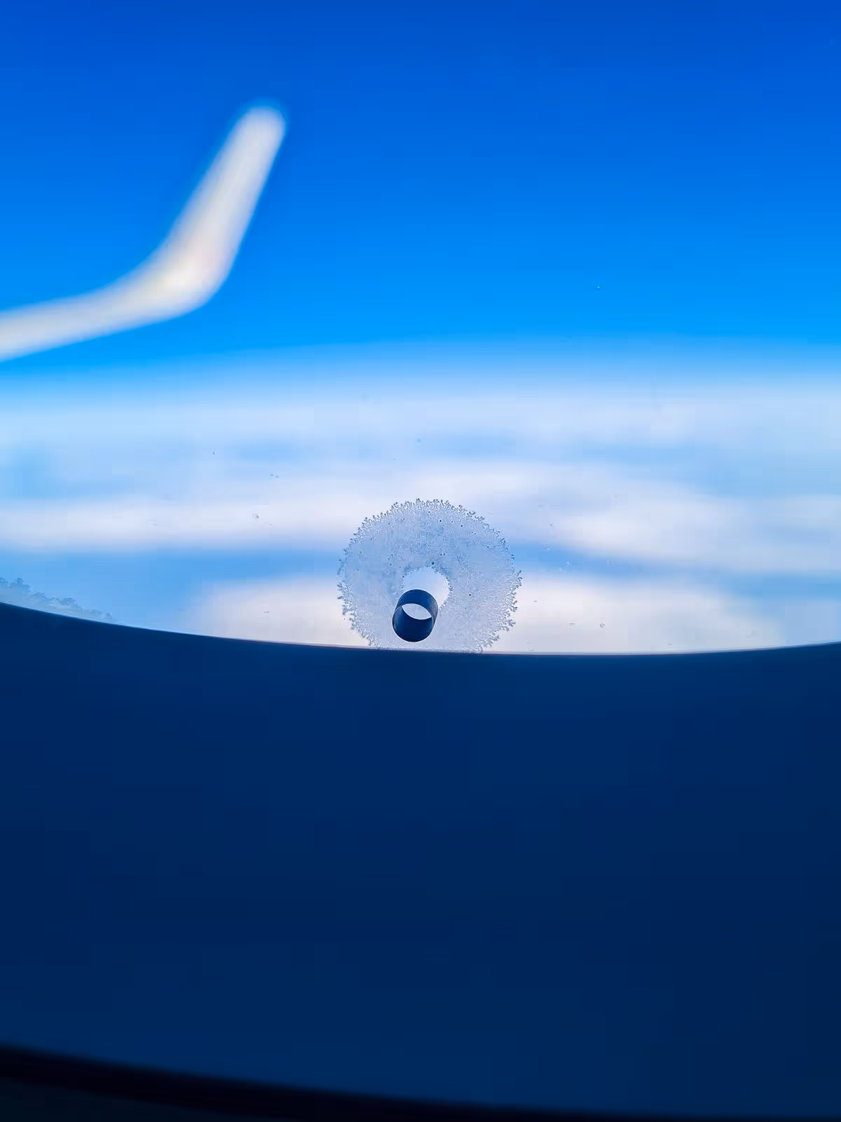 A close-up of a pressure equalization hole with frost forming around it, against a background of an airplane wing, blue sky and clouds. Ideal for travel, aviation, technology or flight themes.