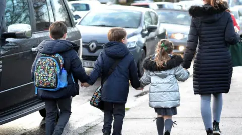 Adult and three schoolchildren with bags hold hands as they walk along the footpath with cars close by on the road. 