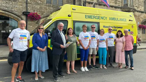 A group of 10 speople stand in a line outside an ambulance smiling at the camera. Some wear white T-shirts with the Ukraine and Union Jack flags on them.