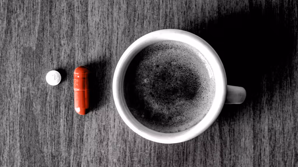 A mug of coffee on a table with a red lozenge and white pill next to it.